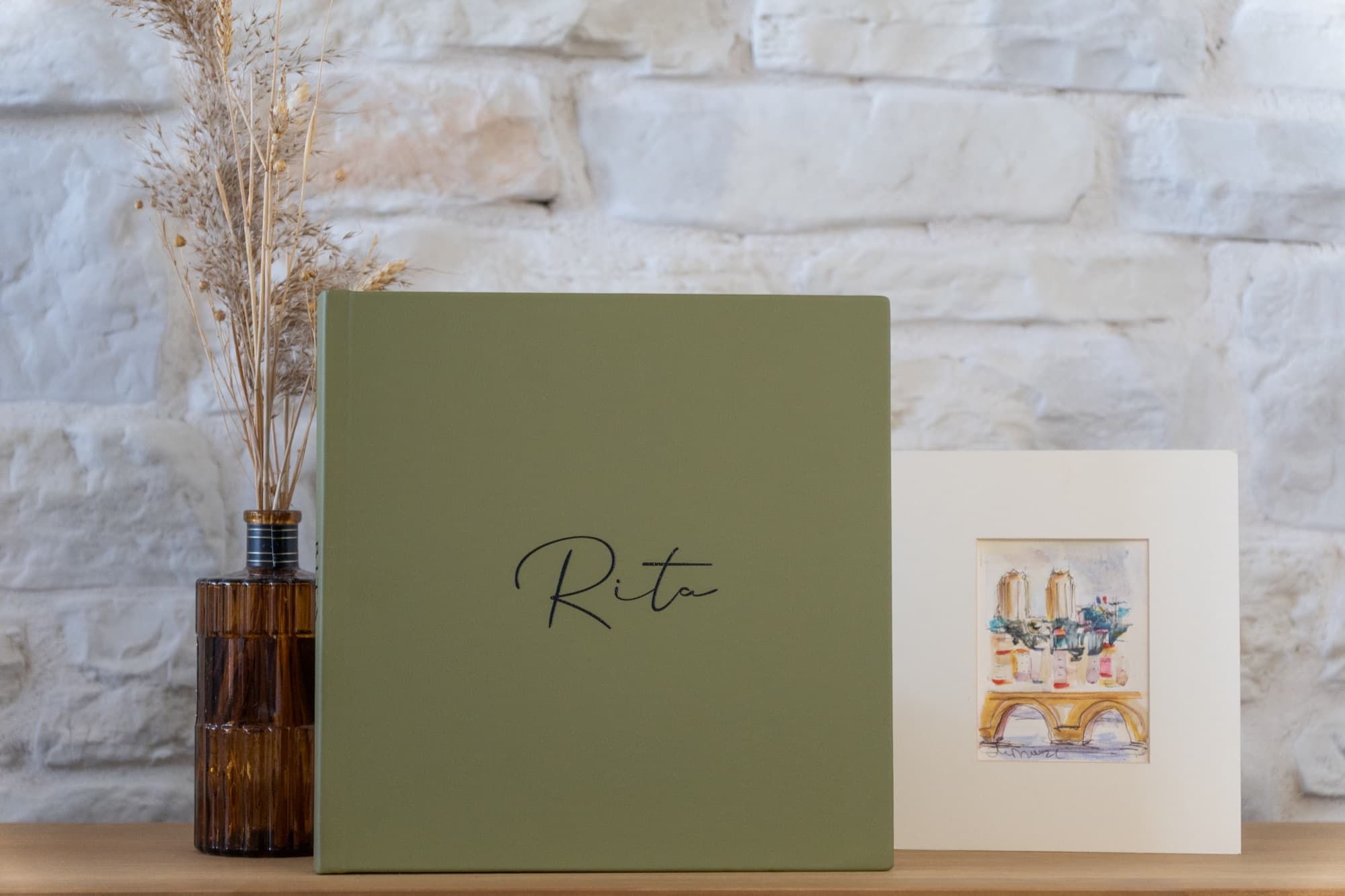A sage green memory book with a name written on the cover, standing against a whitewashed stone wall.